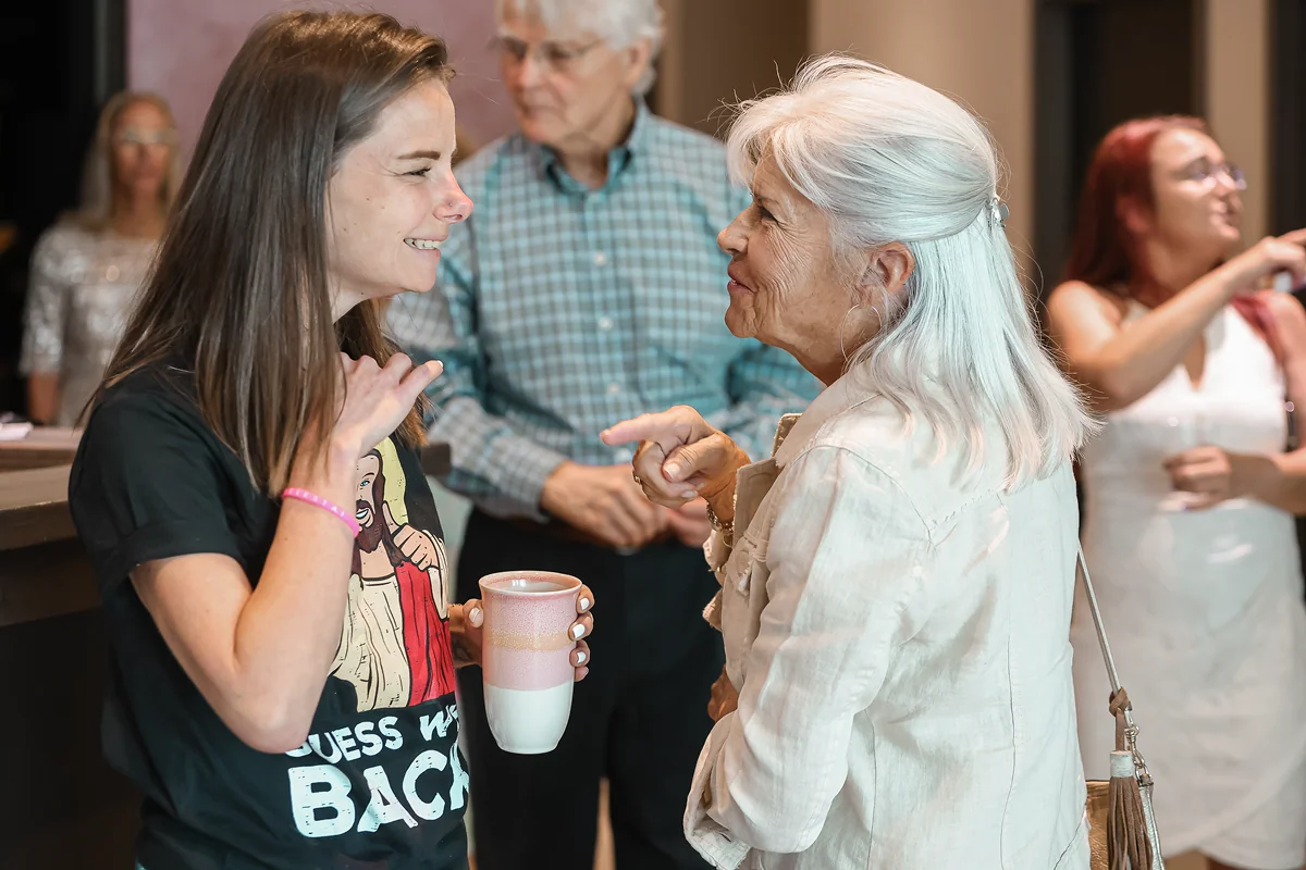 Two women engaged in friendly conversation, one holding a mug, in a social setting with others backgrounded.