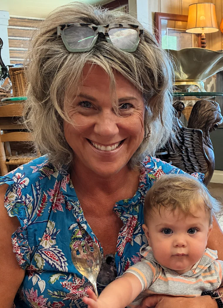 Smiling woman with glasses on her head holding a baby in a colorful shirt indoors.