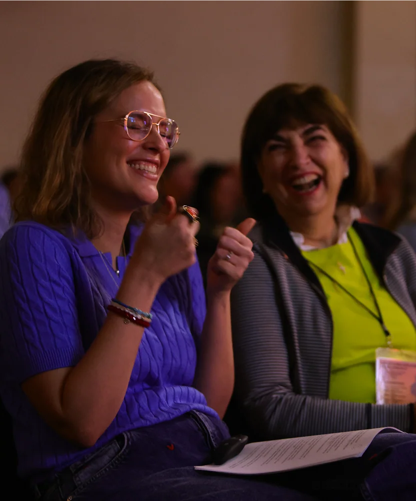 Two women sitting, smiling, and laughing together, one giving a thumbs-up gesture indoors.