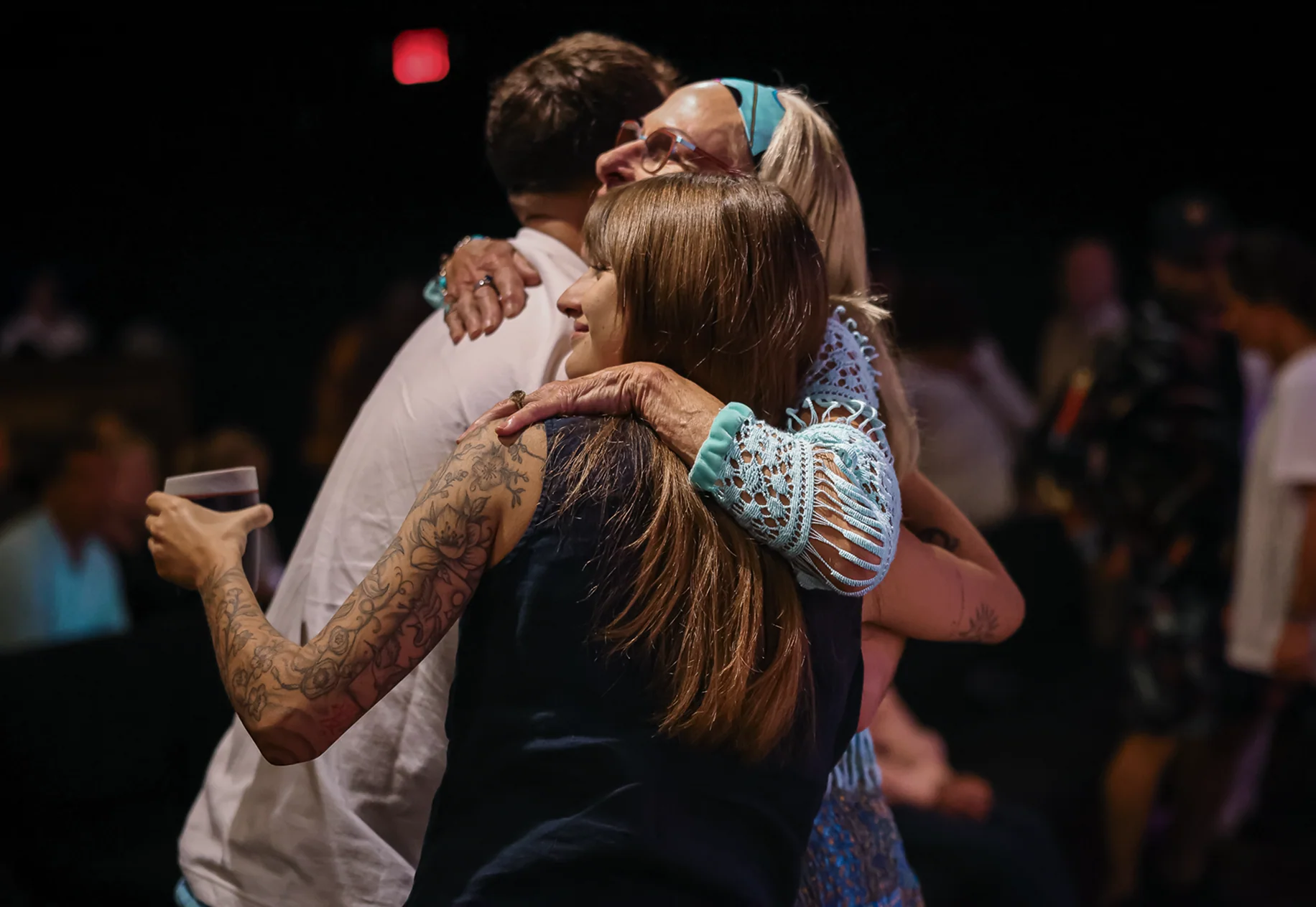 Three people embracing warmly at an event, one with tattooed arm holding a drink.