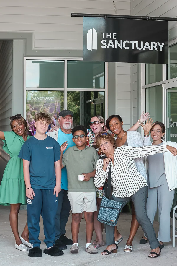 A diverse group of smiling people posing joyfully outside The Sanctuary entrance.