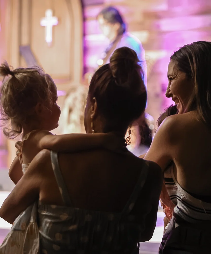 Two women holding children at a church service with a cross and musician in the background.