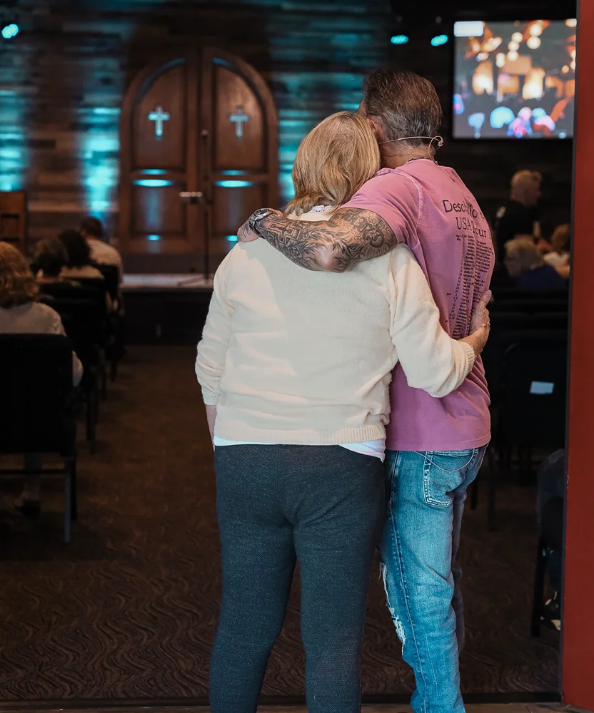 Couple embracing inside a church with wooden doors and crosses in the background during a service.
