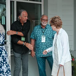 Two men smile and greet a woman at a doorway during a friendly visit.