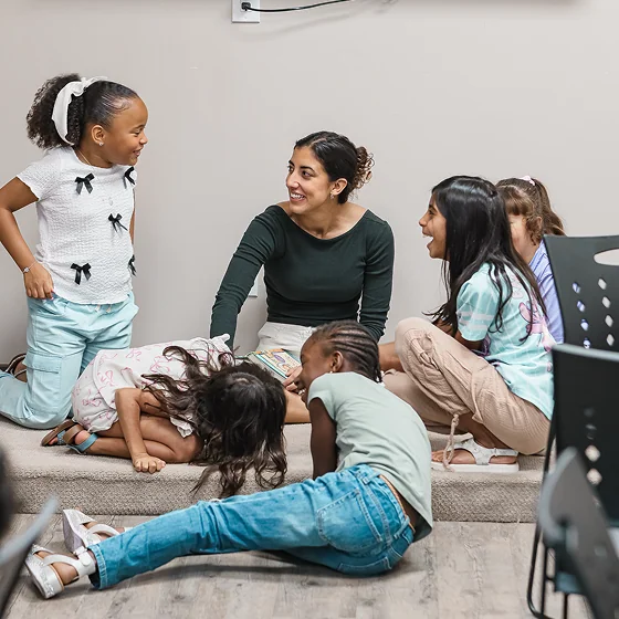 Teacher sitting on floor smiling and interacting with a group of five laughing children indoors.