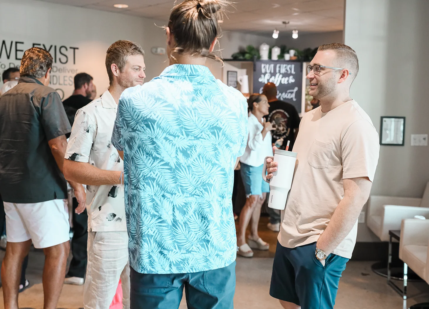 Group of casually dressed men conversing indoors at a social event, one holding a large white cup.