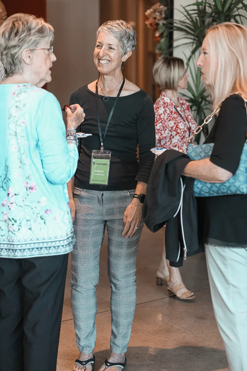 Three women standing and talking indoors at a social event, one wearing a nametag with 'Hilary'.