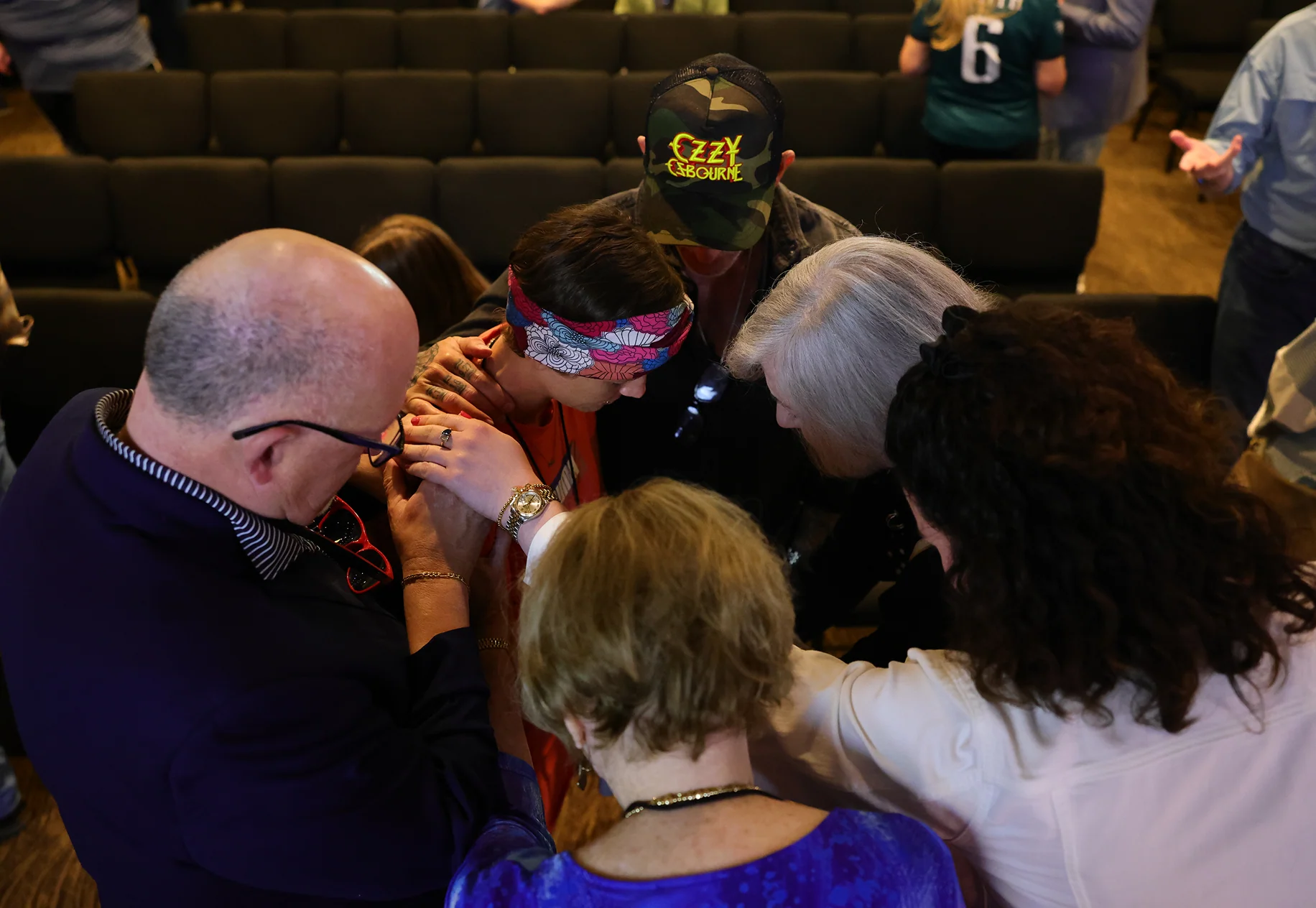 Group of people with heads bowed and hands on a young person in a church seating area.