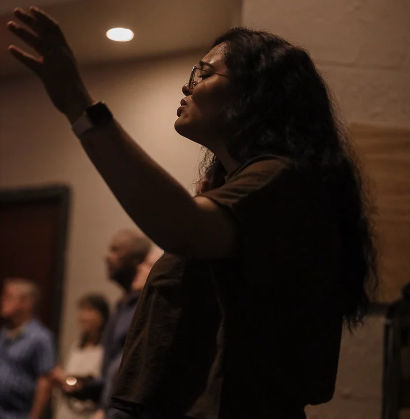 Woman with glasses raising her hand in worship, eyes closed, in a dimly lit room with others blurred behind.