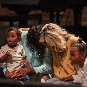 A family sits closely together indoors, heads bowed and hands held in a moment of prayer.