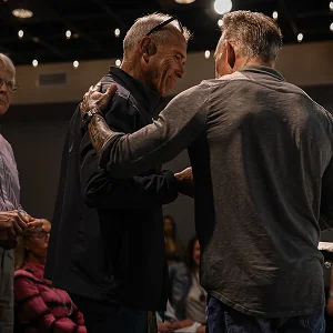 Two men warmly embracing each other indoors with seated people in the background.