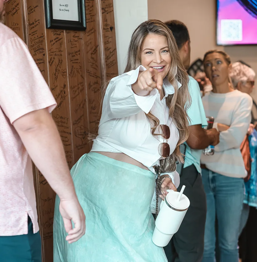 Smiling woman in white shirt and teal skirt pointing at the camera while holding a white tumbler.