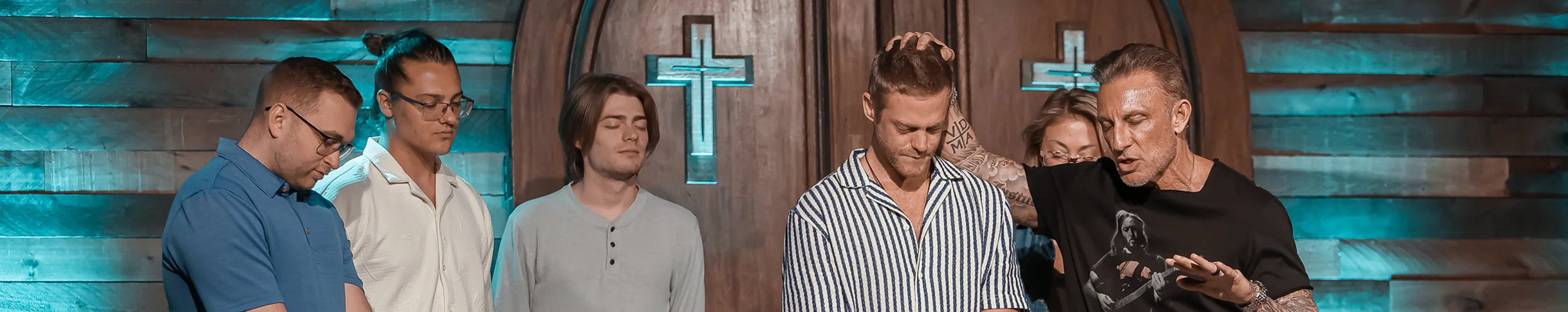 Group of men standing in a church with crosses, one man praying with eyes closed.