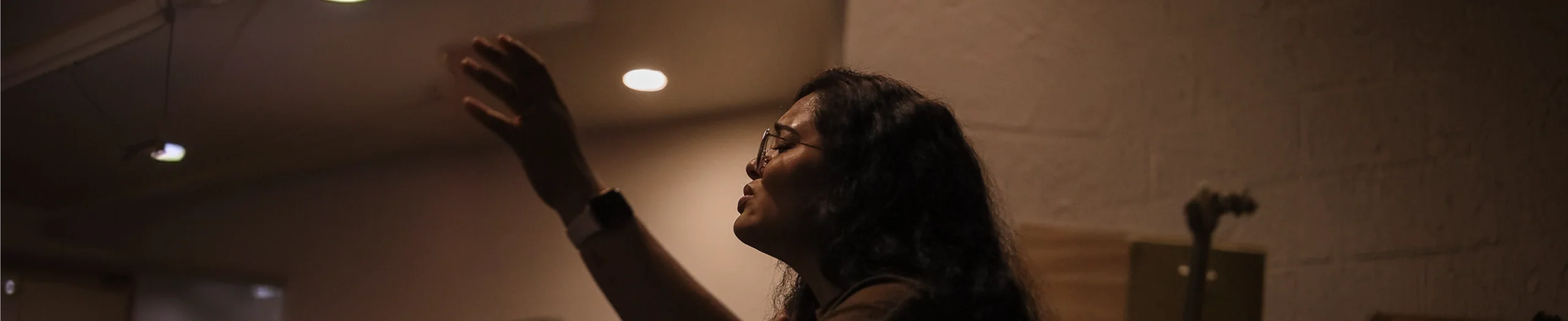 Woman with long hair and glasses reaching upward in a dimly lit room.