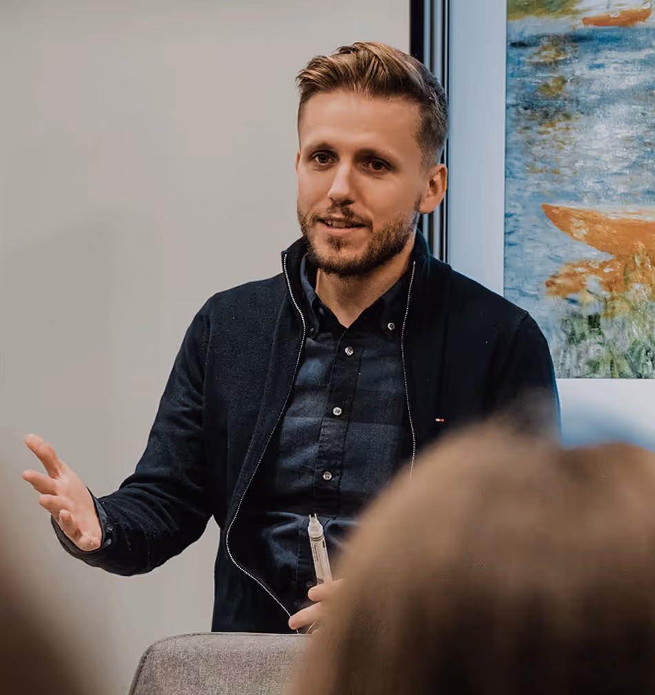A man with light brown hair and a beard is speaking while holding a whiteboard marker, seated in front of a painting and a window.