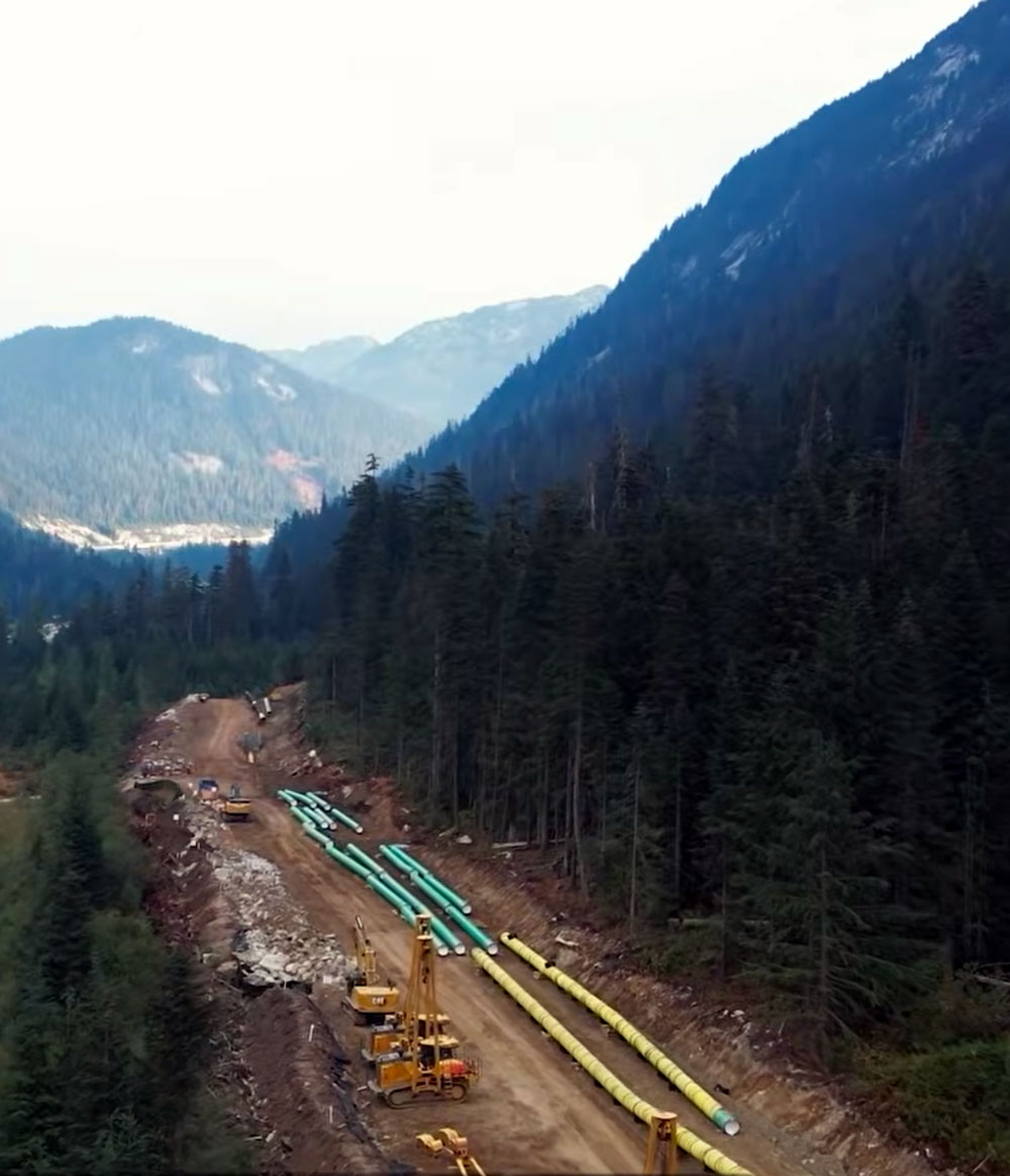 Construction site in a forested mountainous area with large green and yellow pipes laid out along a dirt road, surrounded by evergreen trees and mountain peaks in the background.