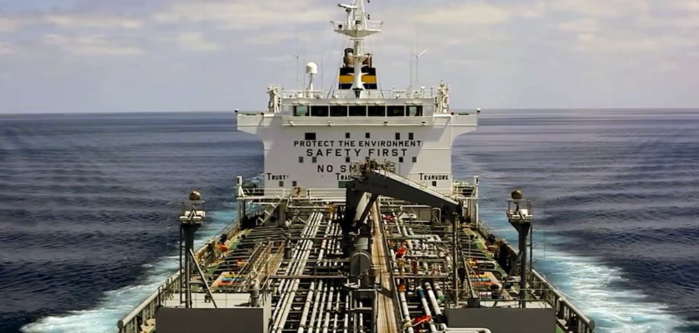 View from the deck of a large tanker ship at sea, showing complex piping and equipment with the bridge in the background displaying safety messages: Protect the environment, Safety first, No smoking.