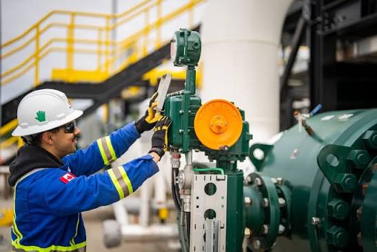 Worker in safety gear including white hard hat and blue protective clothing operates green industrial machinery with orange wheel in a factory setting with yellow stairs in the background.