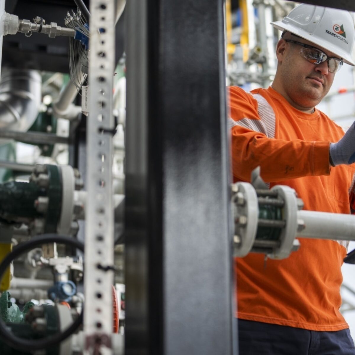 Man wearing an orange safety shirt, white hard hat, and protective glasses working with industrial pipes and equipment.