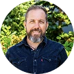 Smiling man with beard and mustache wearing a dark jacket in front of green foliage outdoors.