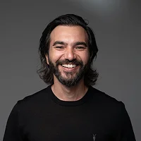 Smiling man with dark hair and beard wearing a black shirt against a gray background.