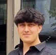 Young man with black hair wearing a black shirt standing outdoors near a building window.