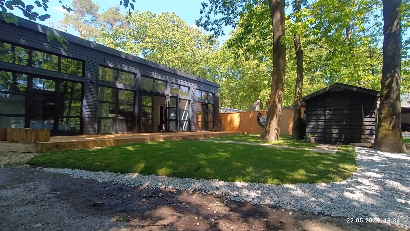 Modern black building with large windows, wooden deck, lawn, trees, and a small black shed.