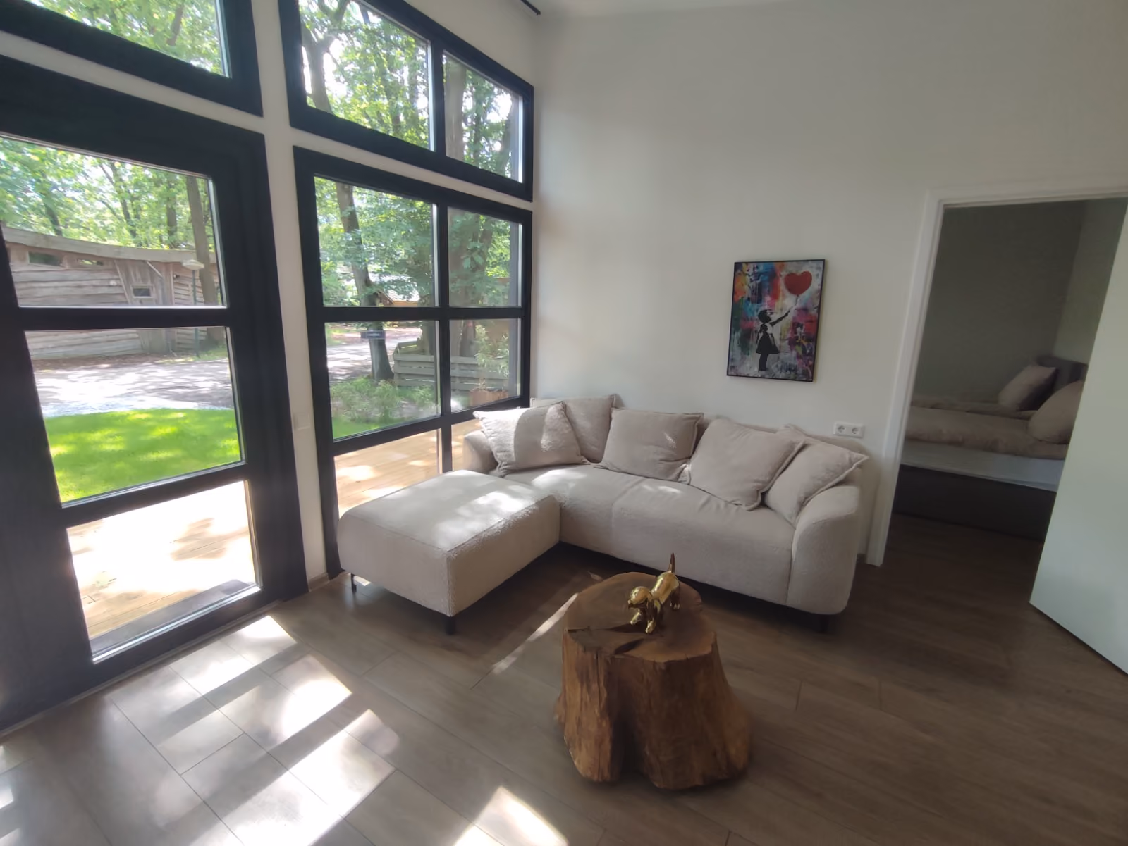 Living room with beige sectional sofa, wooden stump coffee table, large black-framed windows, and artwork on white wall.