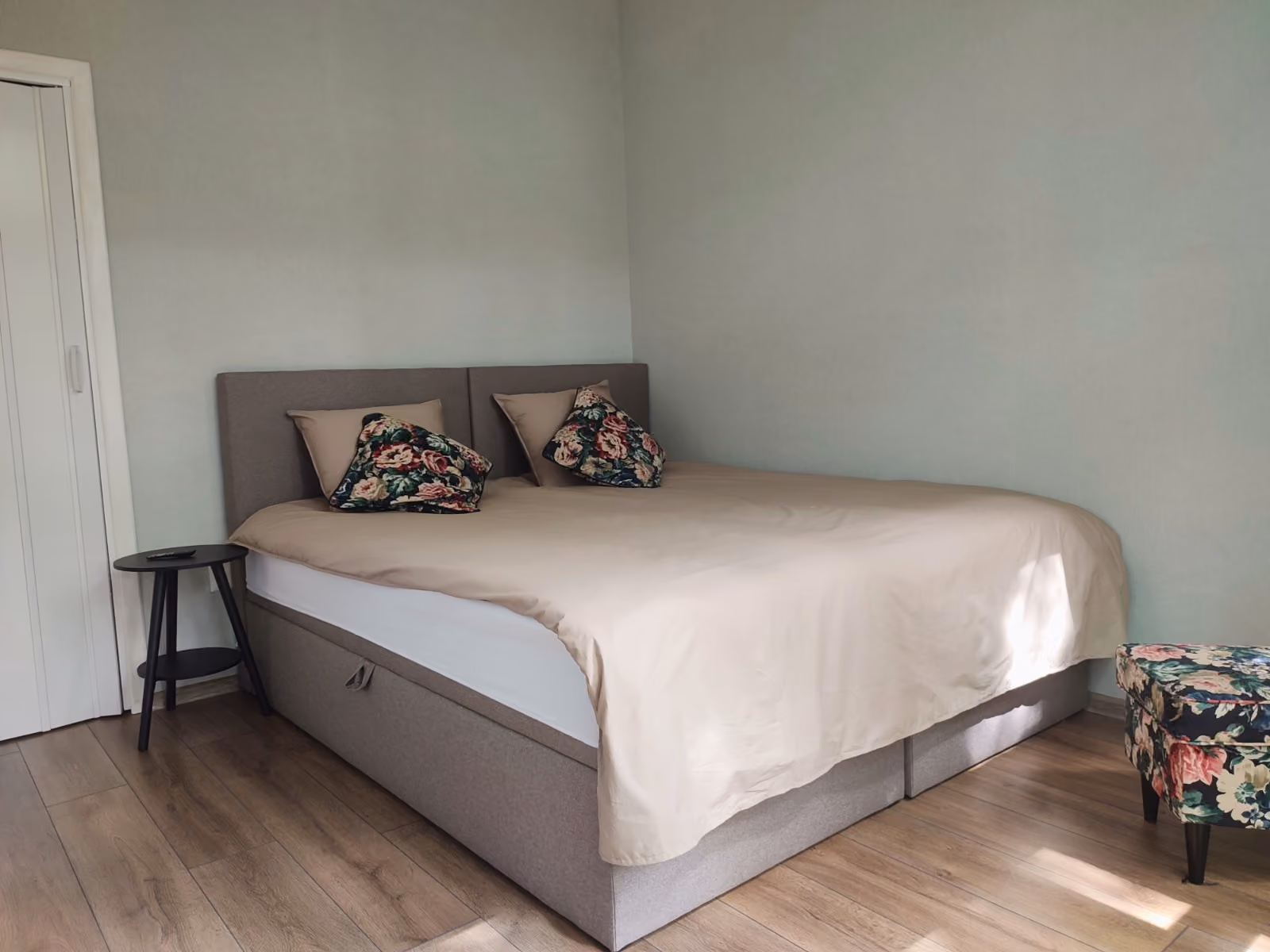 Bed with beige bedding and floral pillows in a room with wooden floor and light wall.