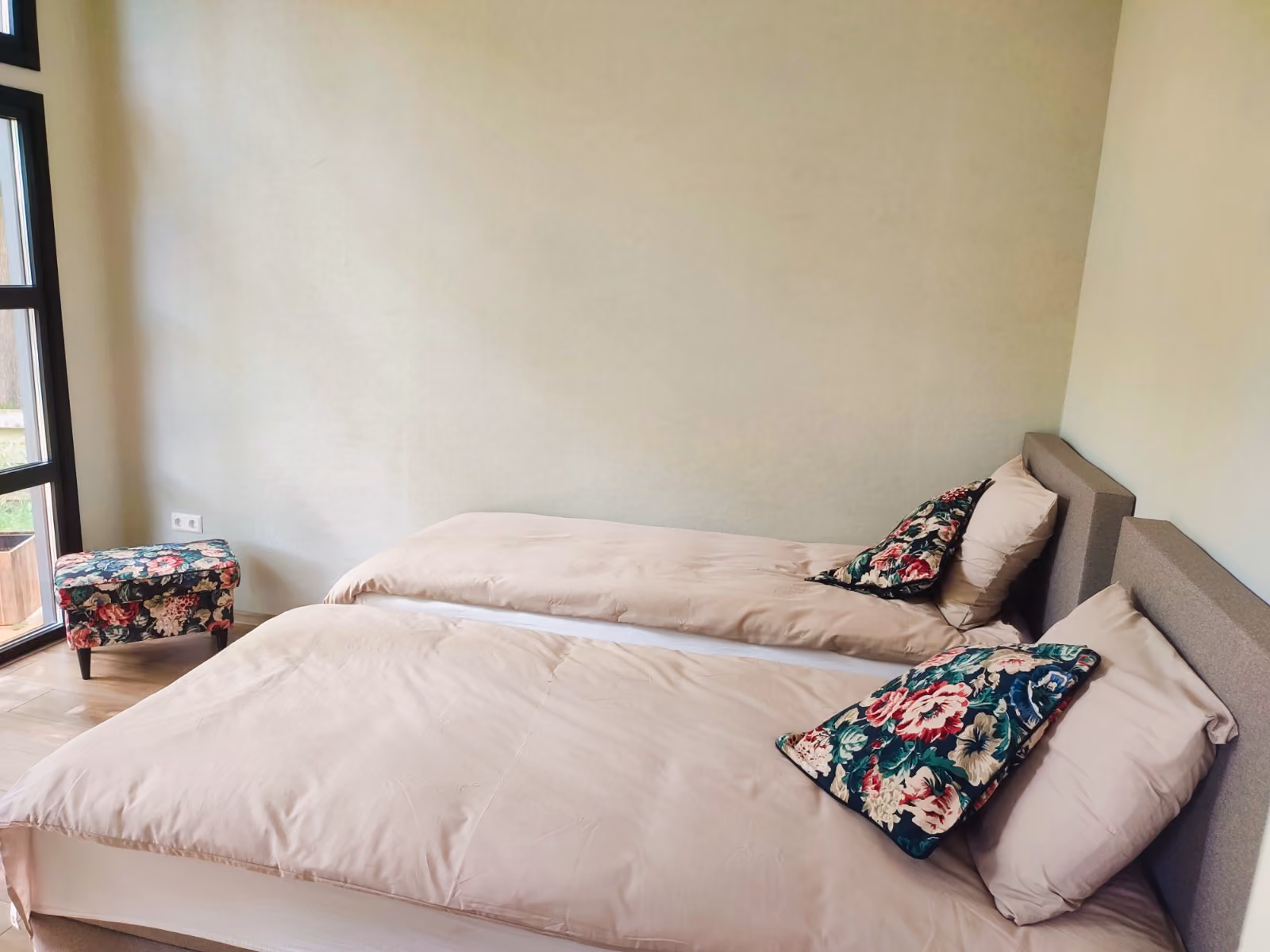 Two beige beds with floral pillows against a plain wall near a window with floral ottoman.