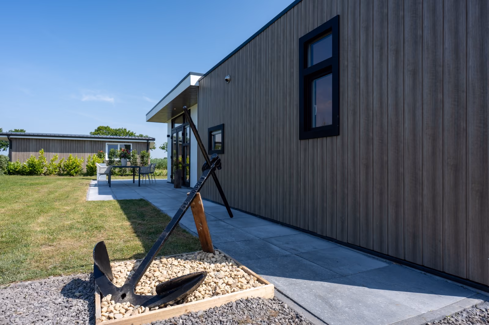Large black ship anchor displayed on stones next to a modern wood-paneled building on a sunny day.