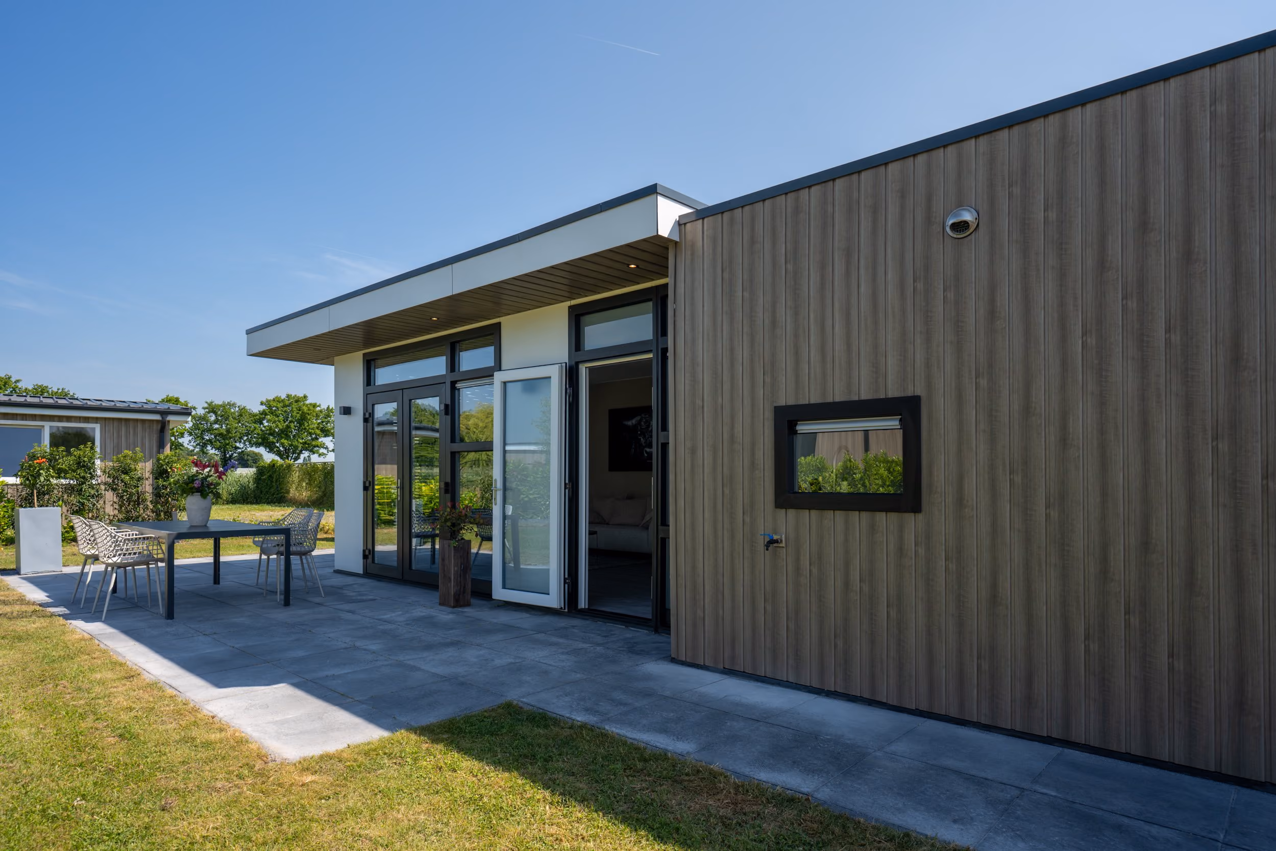 Modern house exterior with wood panel siding, large windows, open door, and patio with table and chairs.