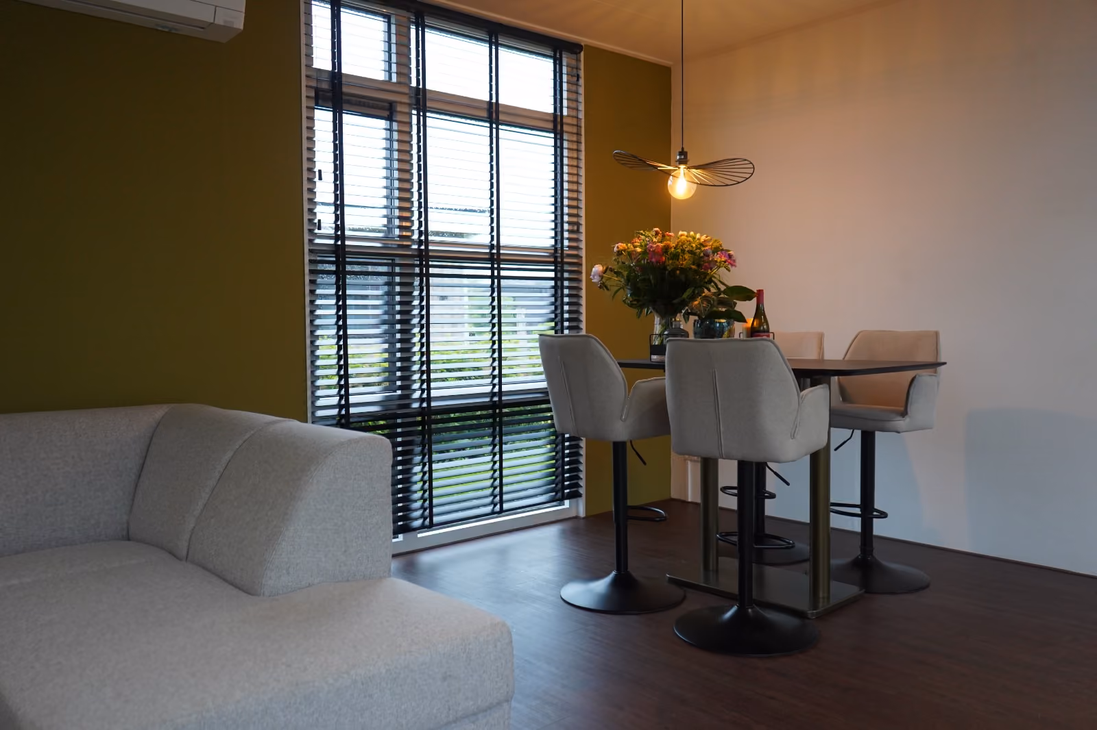 Modern dining area with beige chairs, a black table, flower vase, and large window with blinds.