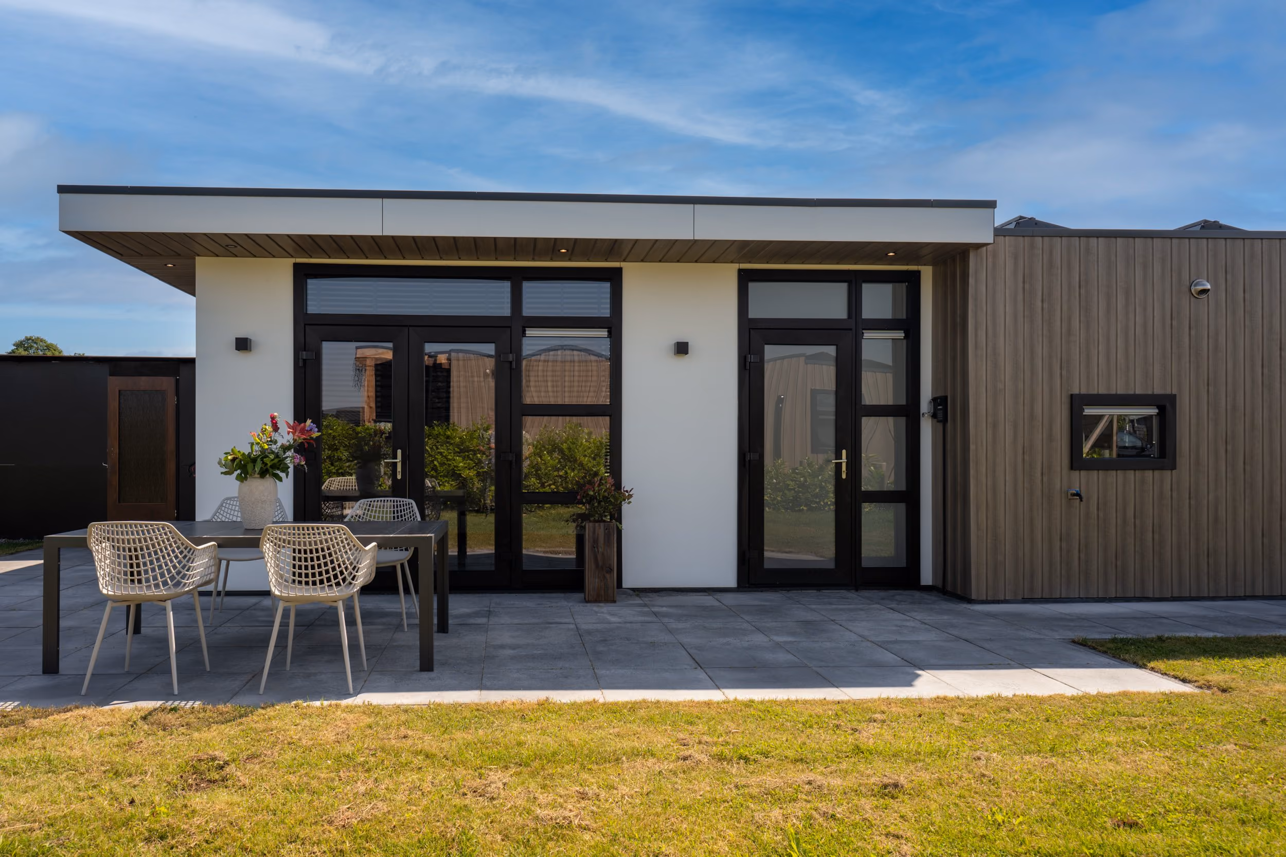 Modern house exterior with black-framed glass doors, outdoor table, and four white chairs on patio.