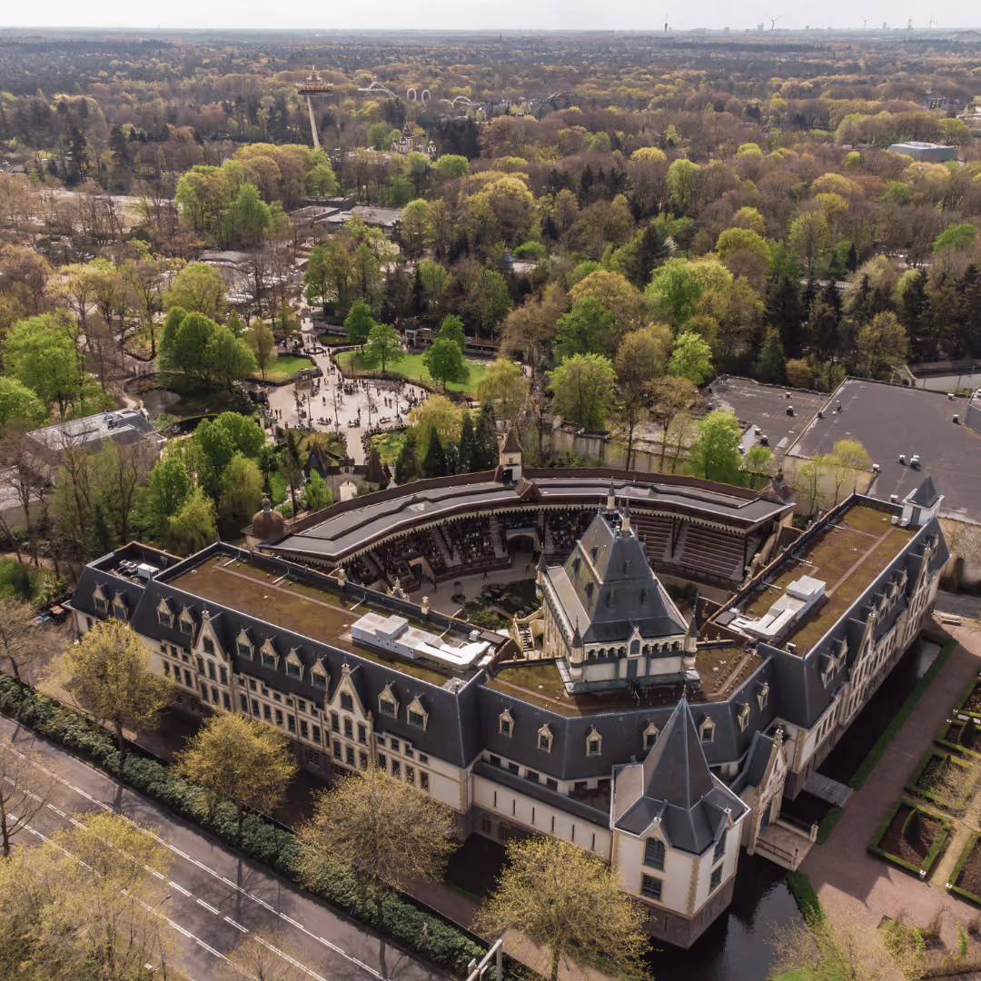 Aerial view of a large castle-like building surrounded by dense green trees and gardens.