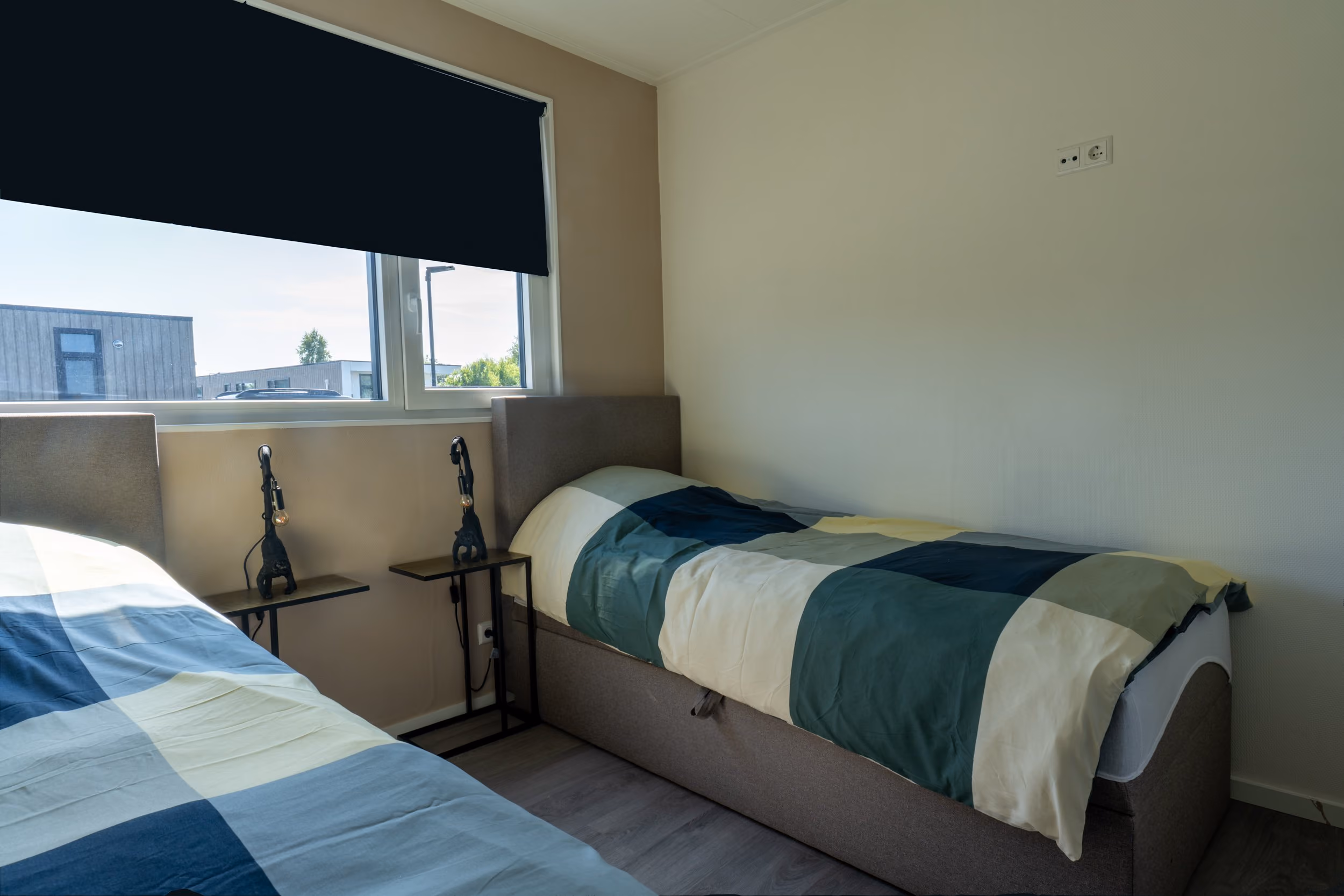 Minimal bedroom with two single beds, patterned duvets, small bedside tables, and a large window with a blind.