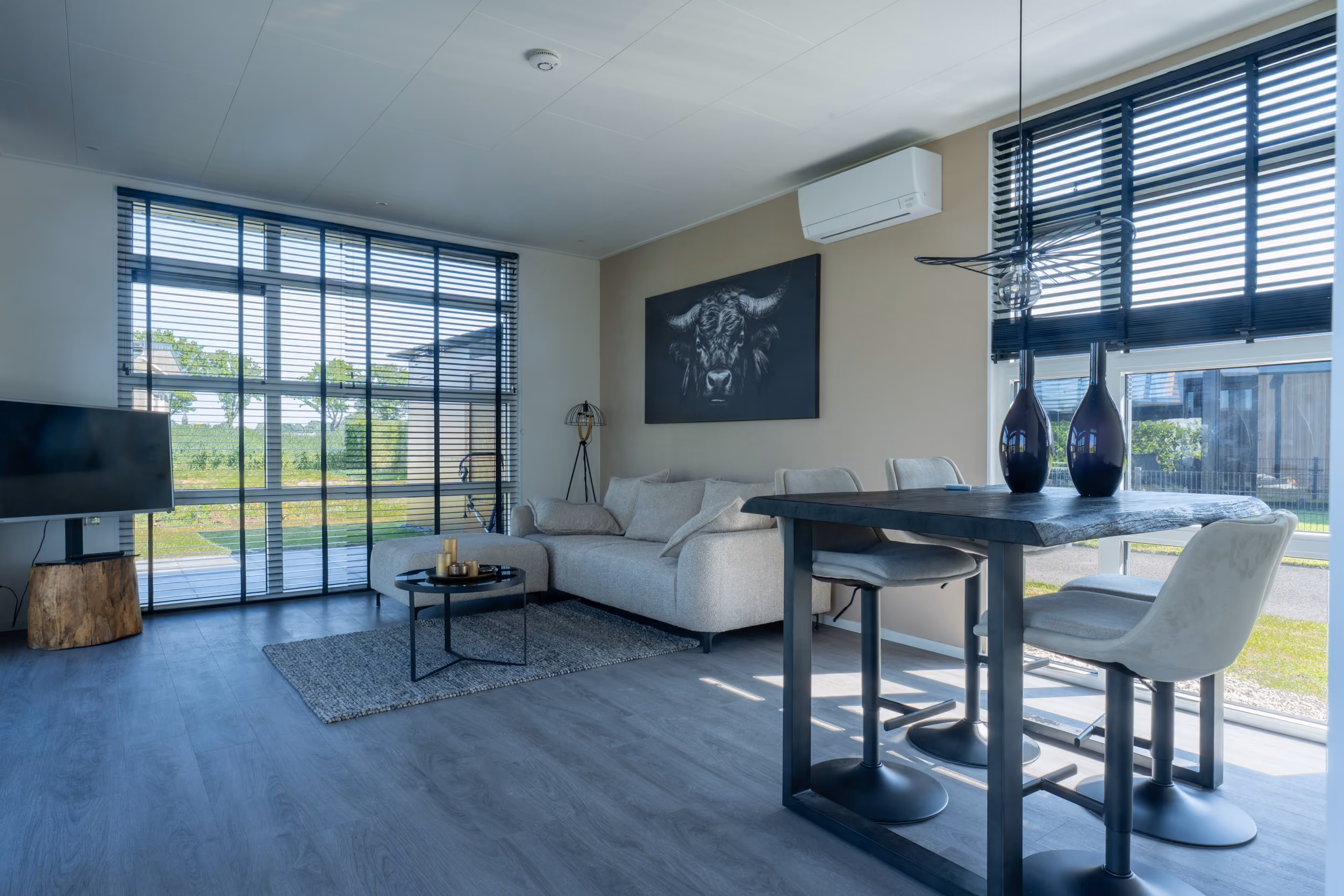 Modern living room with gray sofa, round coffee table, dining table with chairs, and large windows with blinds.