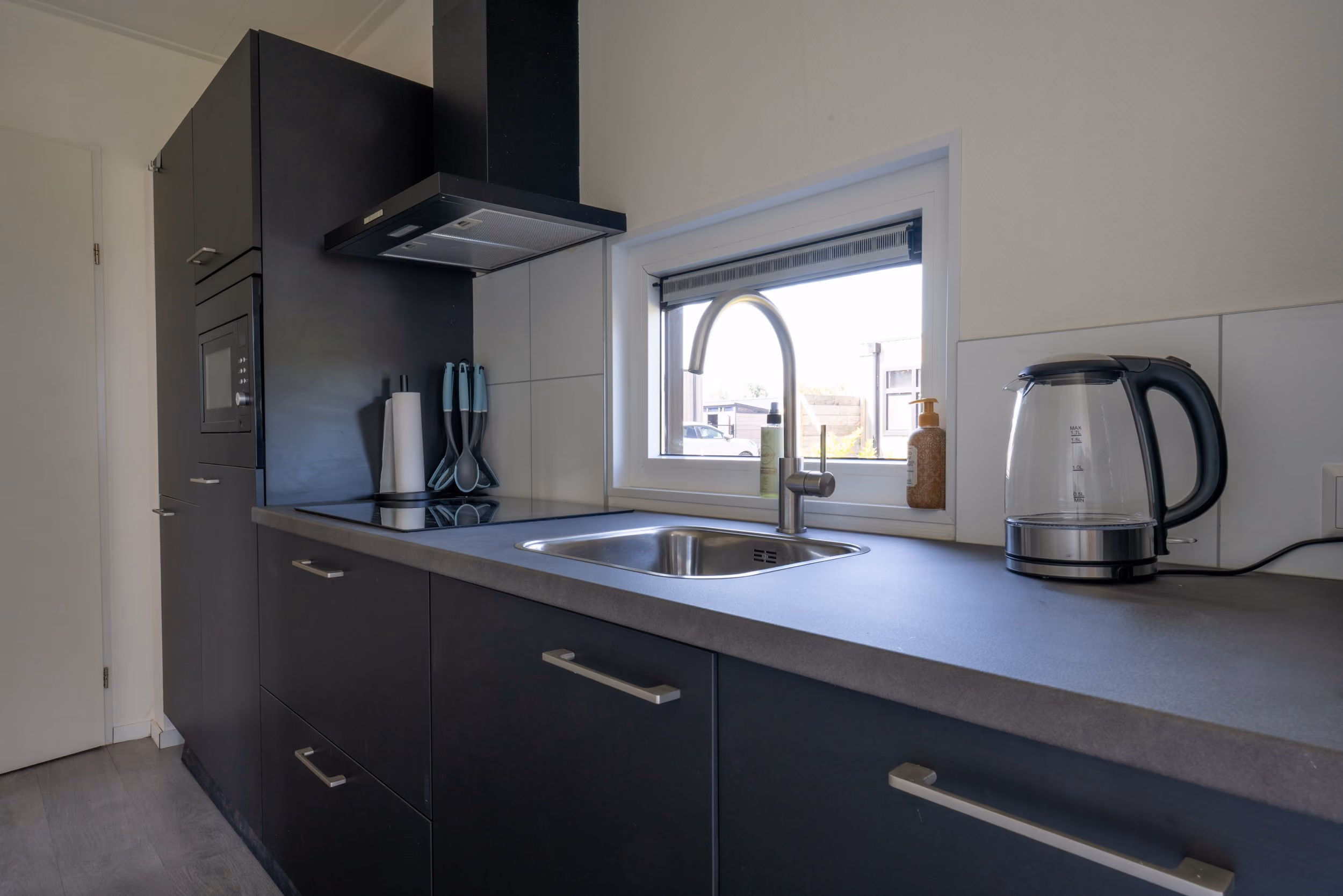 Modern kitchen counter with black cabinets, stainless steel sink, window, and electric kettle.