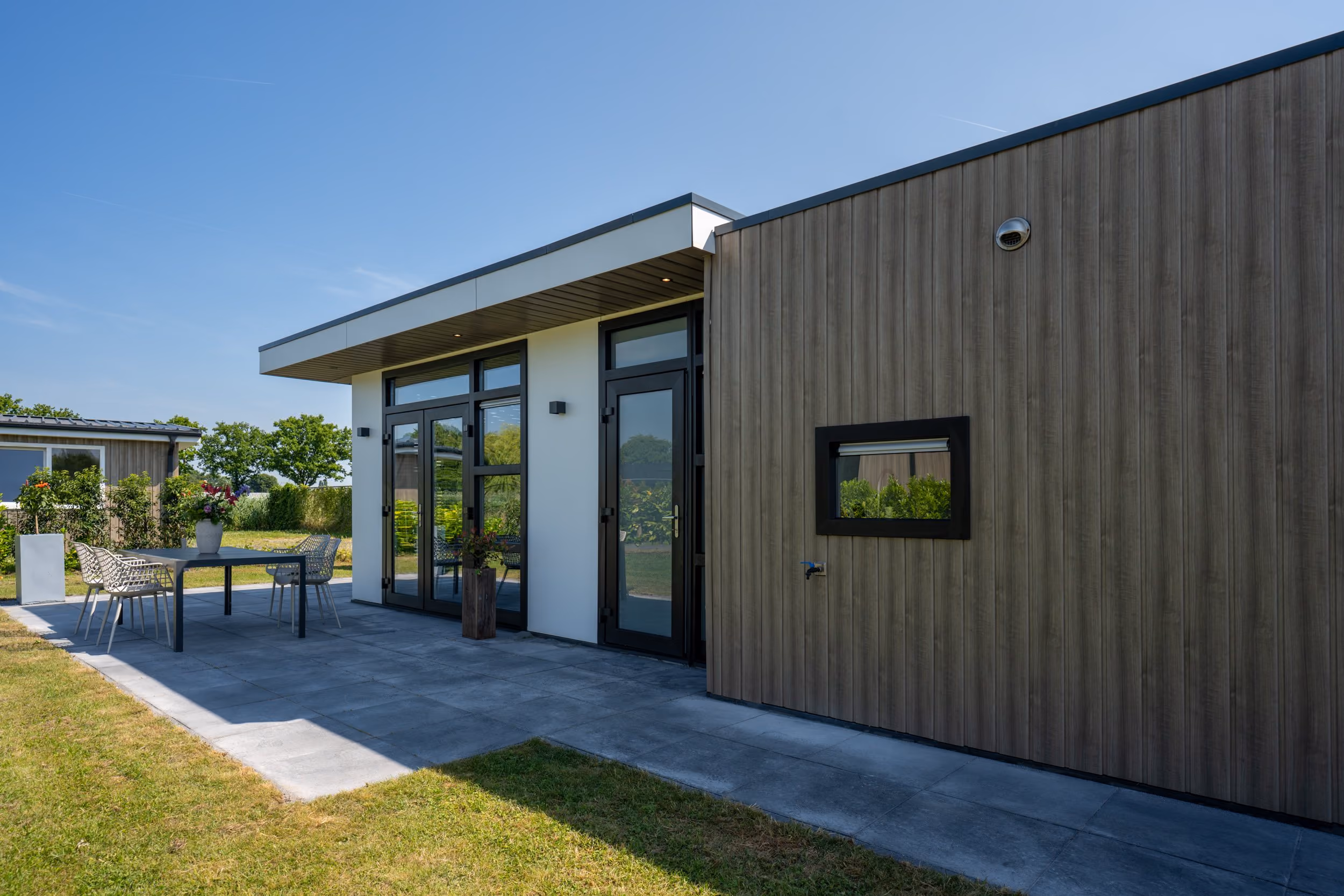 Modern patio with glass door, table, chairs, and wood-paneled house exterior under a clear blue sky.