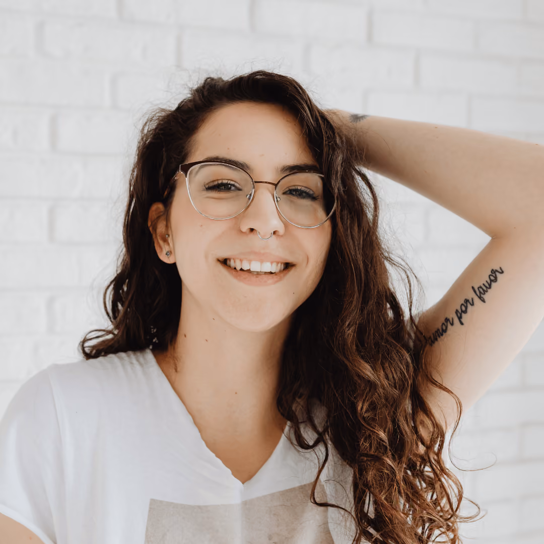 Smiling woman with long curly hair, glasses, and arm tattoo reading 'Amor por favor'.