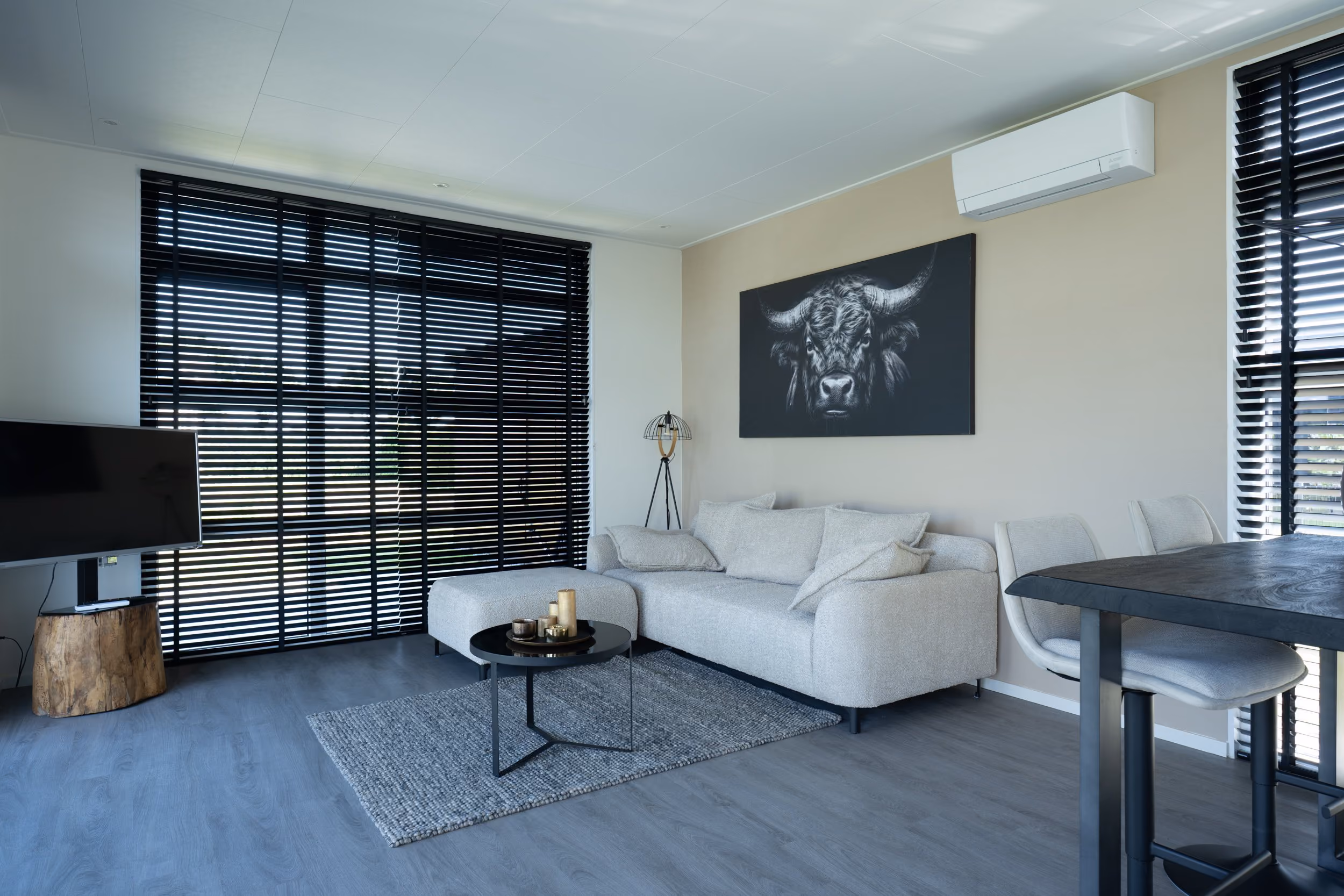Modern living room with gray sofa, black round coffee table, wall art of a bull, and large windows with blinds.