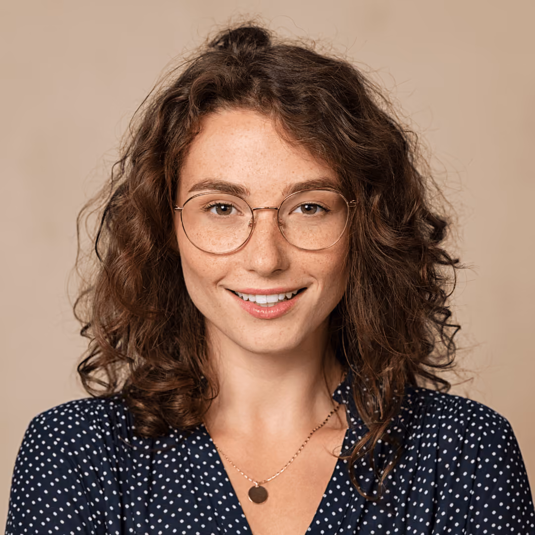 Smiling woman with curly hair, glasses, and a navy polka dot blouse on beige background.