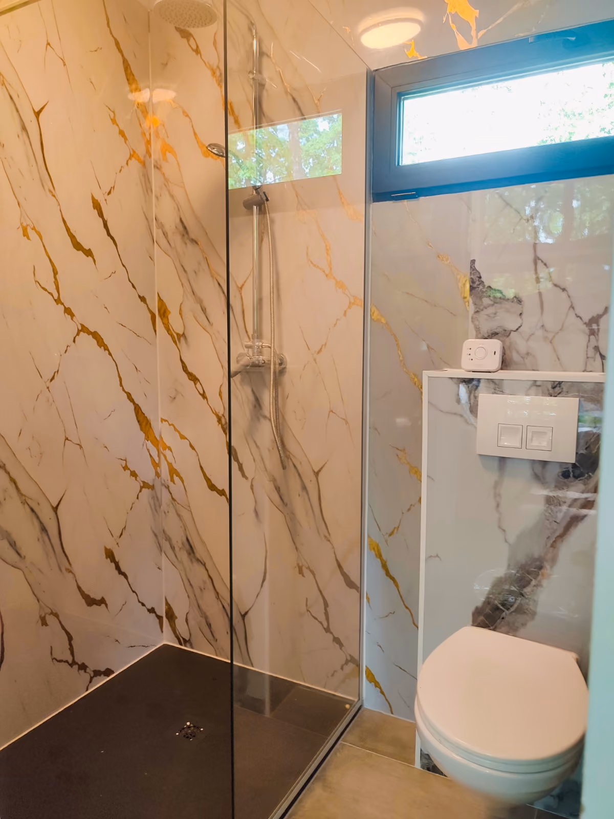 Modern bathroom with marble-patterned walls, glass shower enclosure, and a wall-mounted toilet beneath a small window.