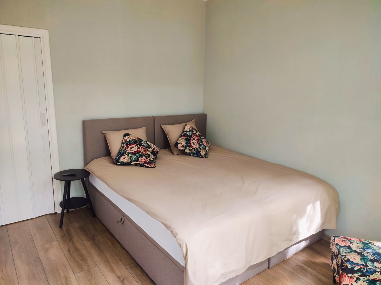 Minimalist bedroom with beige bed, floral pillows, small black side table, and light green walls.