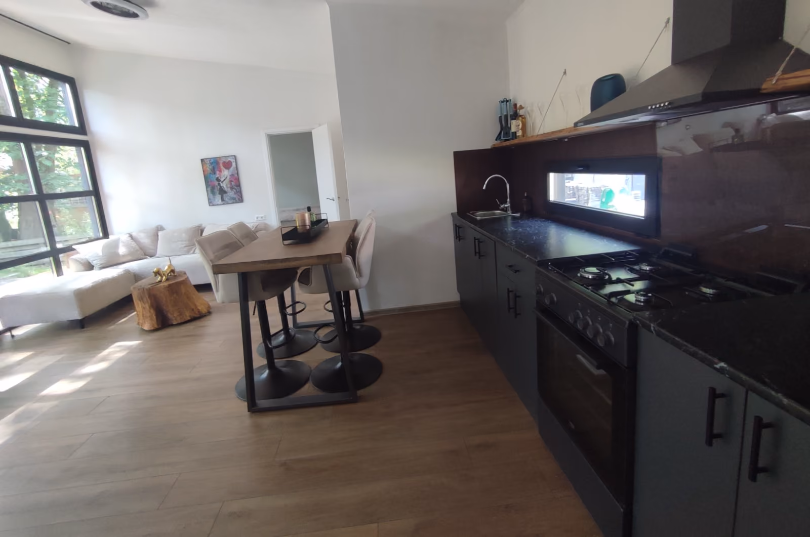 Modern kitchen with black countertops, gas stove, and a wooden dining table near a bright living room.