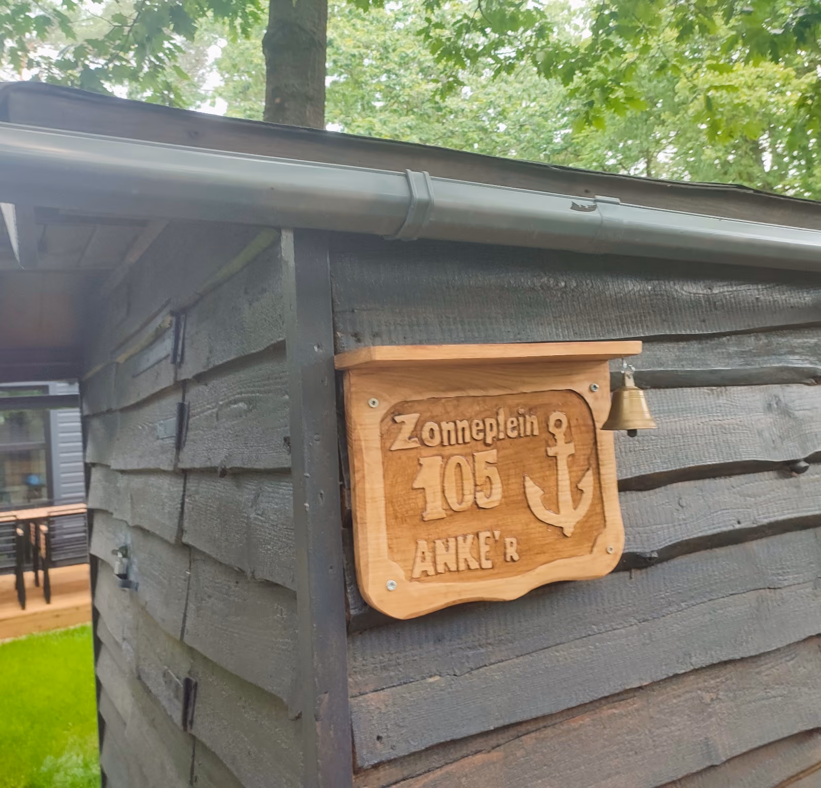 Wooden sign with anchor and text 'Zonneplein 105 ANKER' mounted on dark wood wall with small bell.