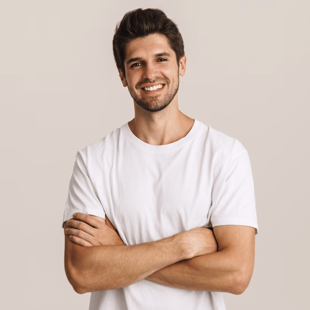 Smiling man with dark hair and beard wearing a white t-shirt, standing with arms crossed against beige background.