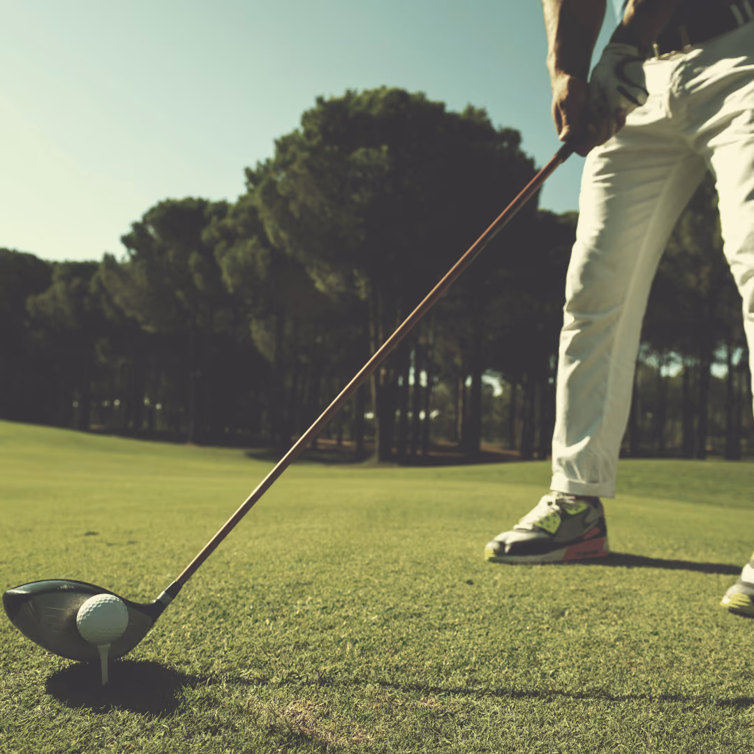 Golfer wearing white pants preparing to hit a golf ball with a driver on a sunny course.