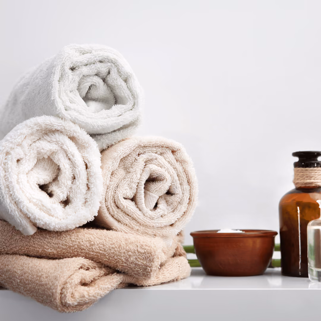 Stack of beige and white rolled towels next to a brown bowl and an amber glass bottle on a white surface.