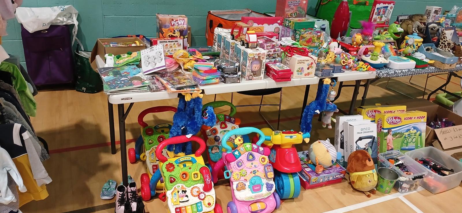 Tables filled with various children’s toys including walkers, stuffed animals, books, and boxed games at a sale.